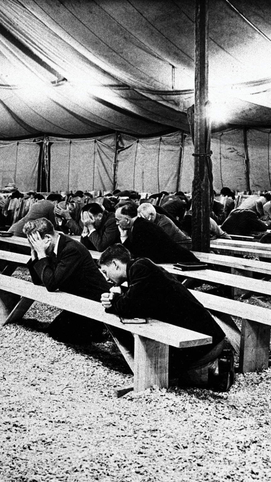 People praying inside a large tent, sitting on long benches, with a focused and solemn demeanor.
