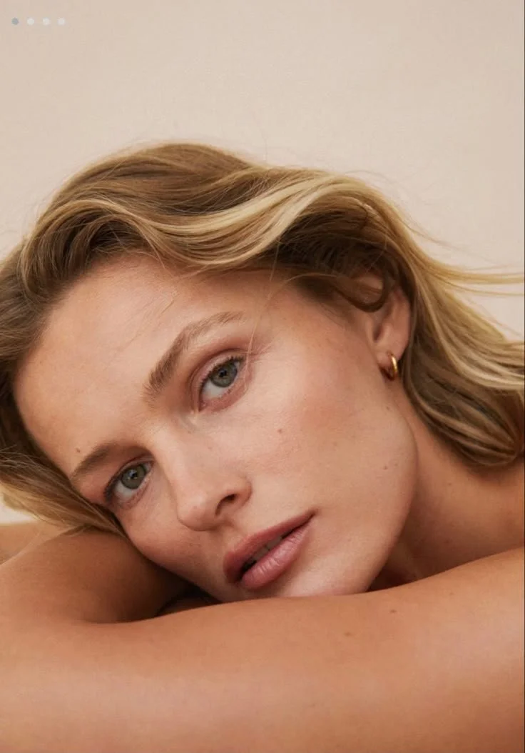 A close-up of a young woman with blonde hair resting her head on her arm, looking at the camera with a soft expression.