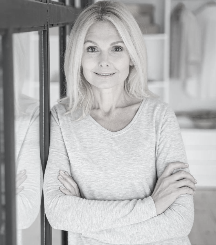 A woman with shoulder-length blonde hair, wearing a light-colored long-sleeve shirt, standing with crossed arms and smiling at the camera in a modern, bright interior space.