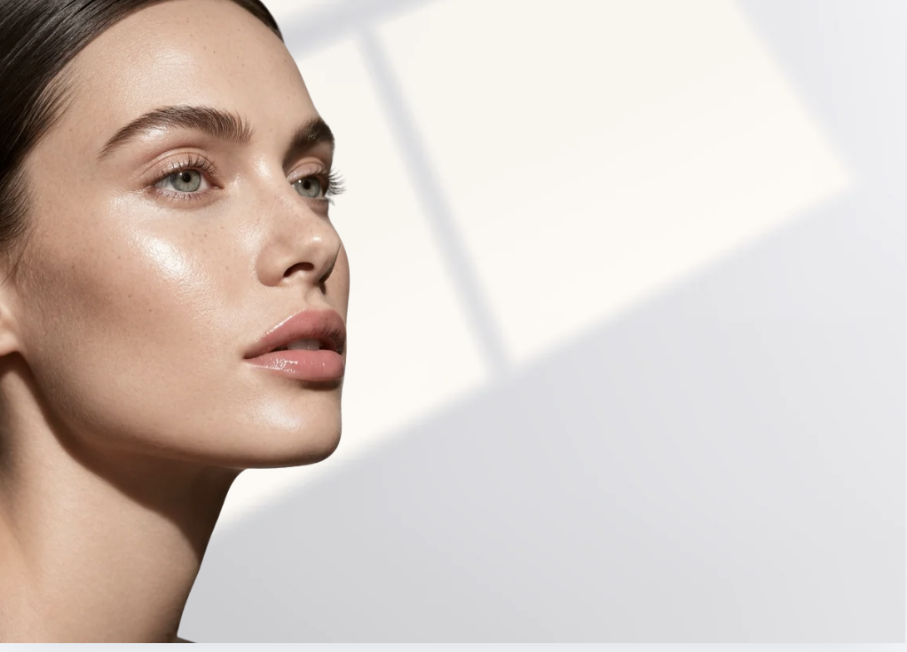 Close-up of a woman with clear skin, light makeup, and light-colored eyes against a plain background.