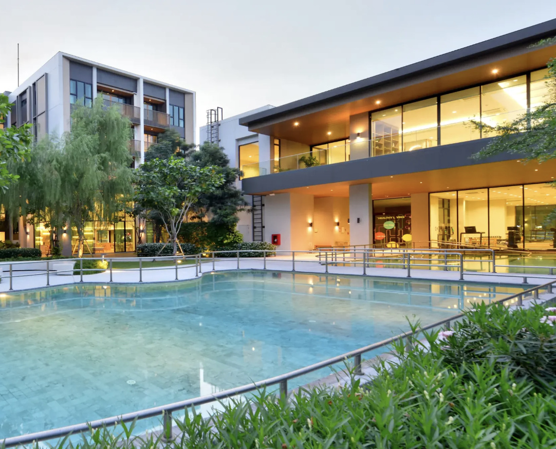 Modern hotel with large swimming pool, surrounded by trees and illuminated interior windows, during early evening.