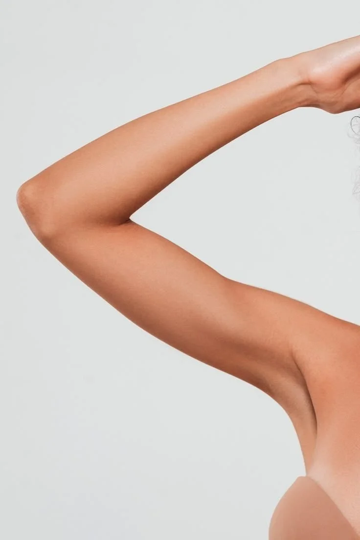 Close-up of a woman's bare arm raised with her hand touching her head, against a plain white background.