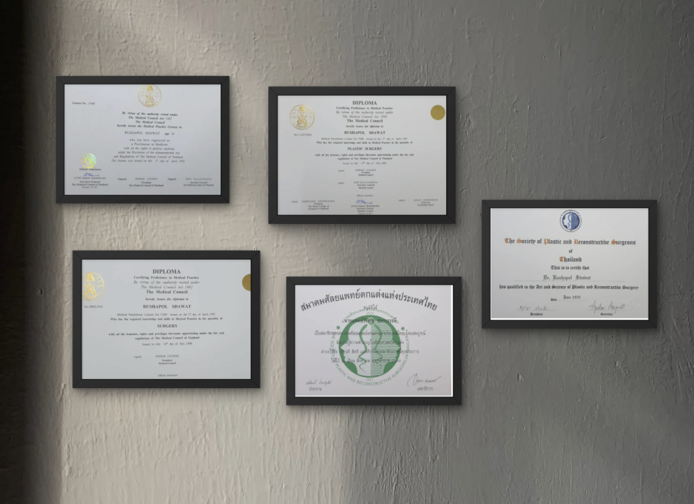 Five framed diplomas and certificates on a wall, with three at the top and two at the bottom, in black frames against a textured beige and gray wall.
