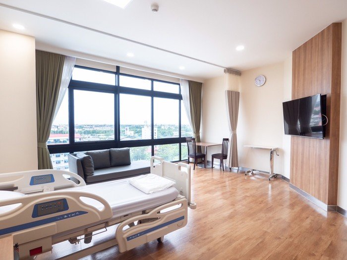 Hospital room with a bed, large window with curtains, a small sofa, a TV mounted on a wooden wall, two chairs near a window, and a clock.