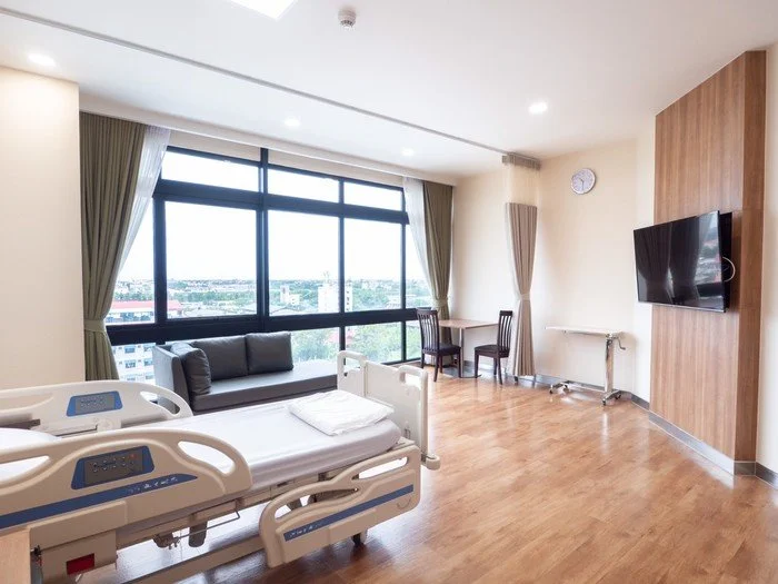 Hospital room with a hospital bed, a gray sofa, a large window with a city view, two chairs near a small table, a wall-mounted TV, and a wooden accent wall.