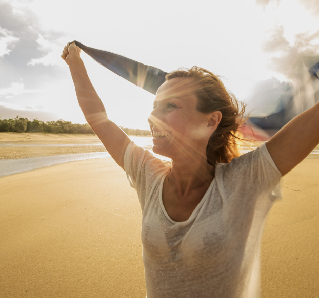 A woman celebrating joyfully on a beach with her arms raised, holding a jacket or shirt, with sunlight shining through the clouds.