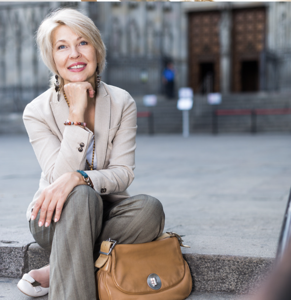 Smiling woman with blonde hair sitting on sidewalk with a brown handbag, outside building with wooden door in background.