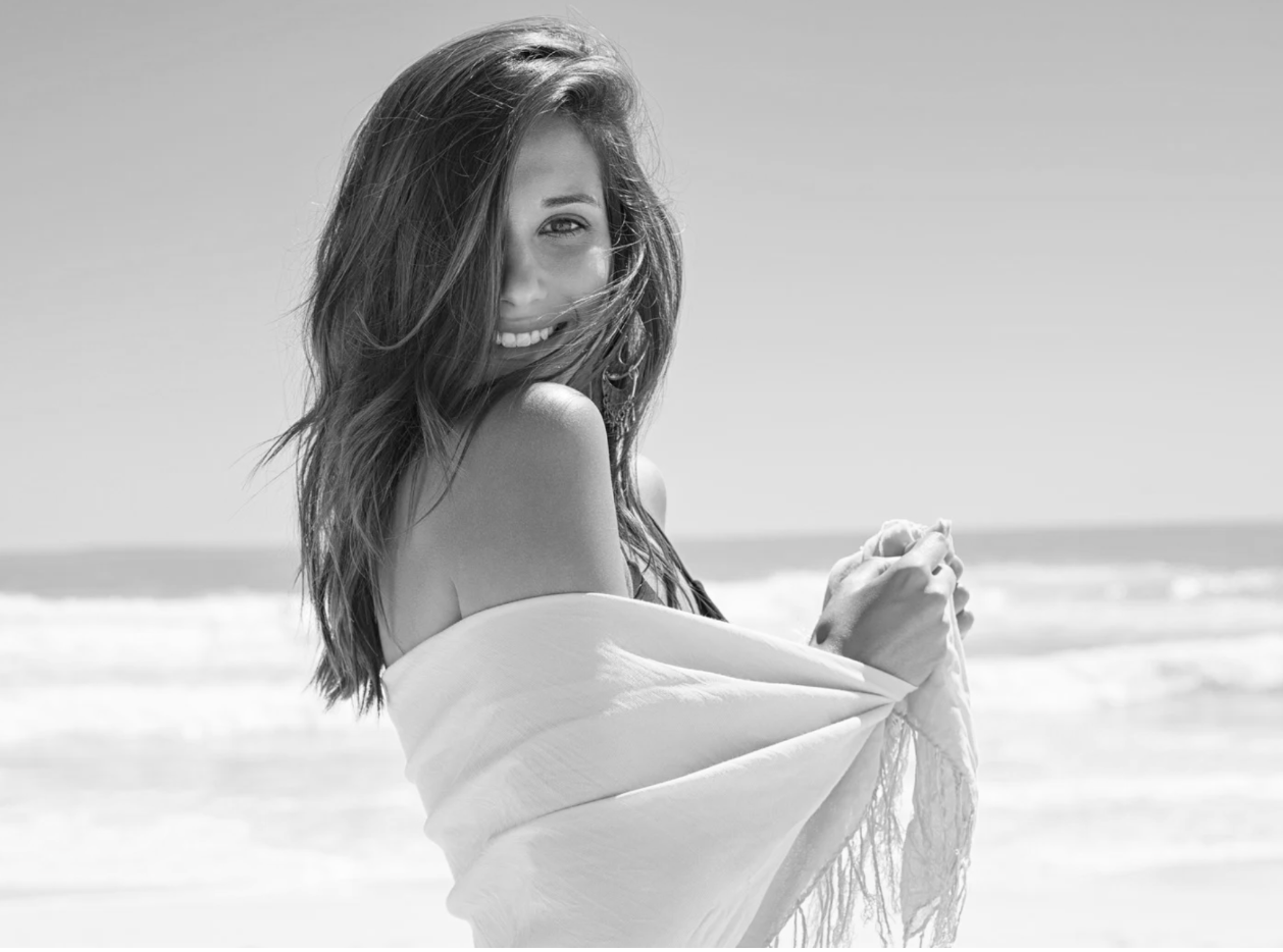 A smiling woman with long hair on a beach, holding a towel, in black and white.