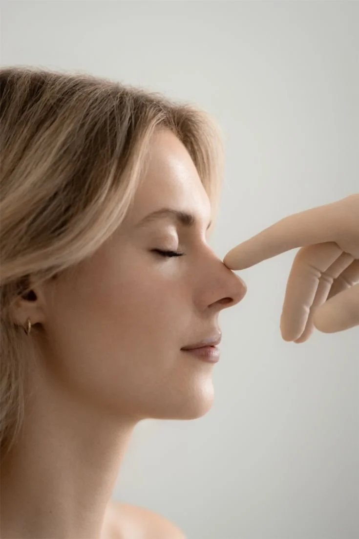 A woman with closed eyes and blonde hair has her nose gently pressed by a gloved finger.