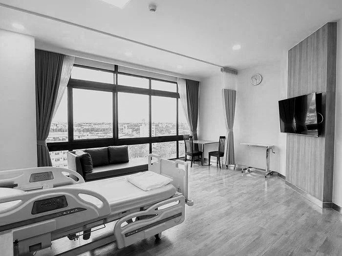 A hospital room with a bed, large window with curtains, a sofa, two chairs, a wall-mounted TV, a clock, and a small table.