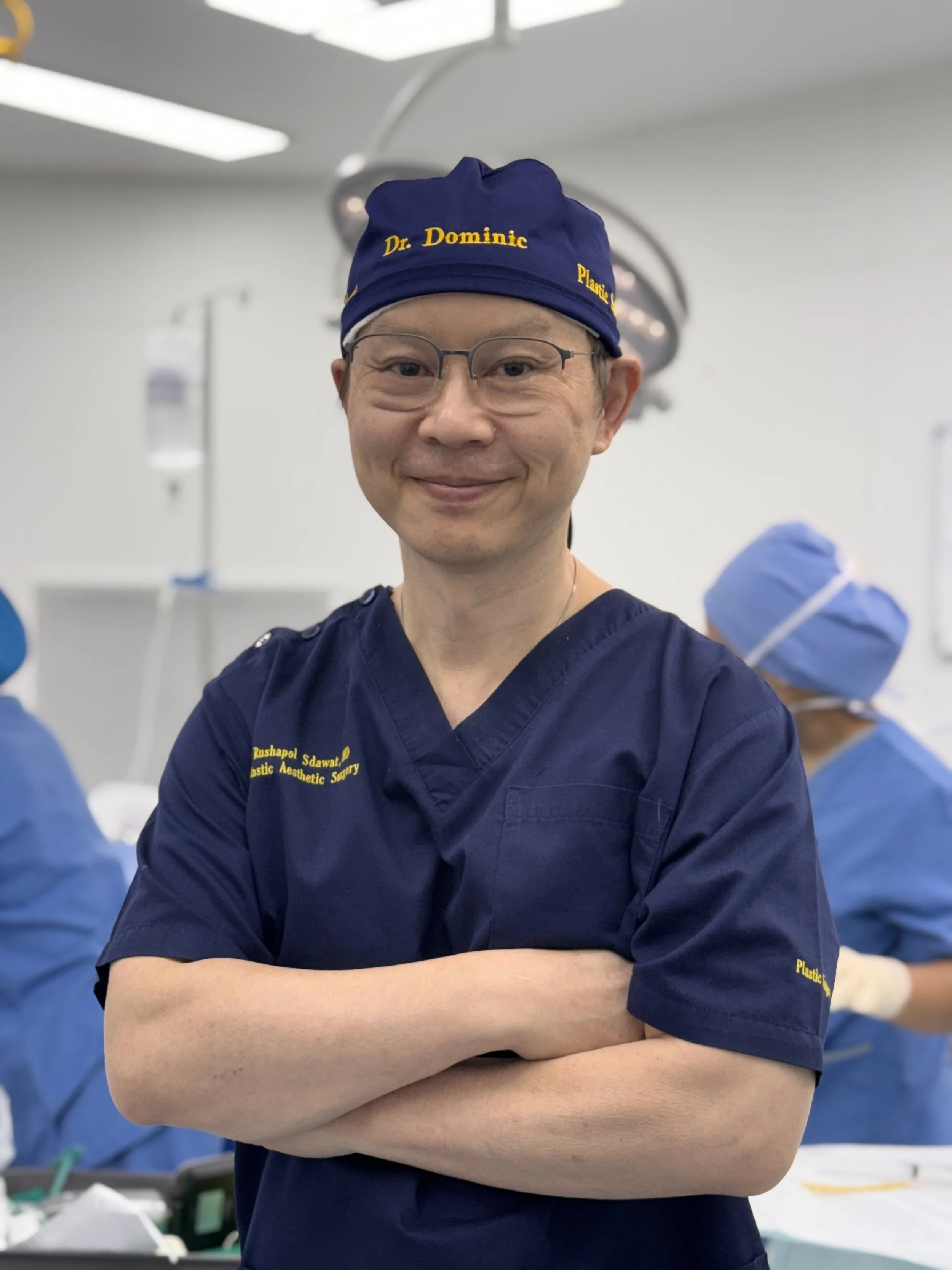 A smiling male surgeon in blue scrubs, glasses, and a blue surgical cap with yellow text, standing with arms crossed in an operating room.