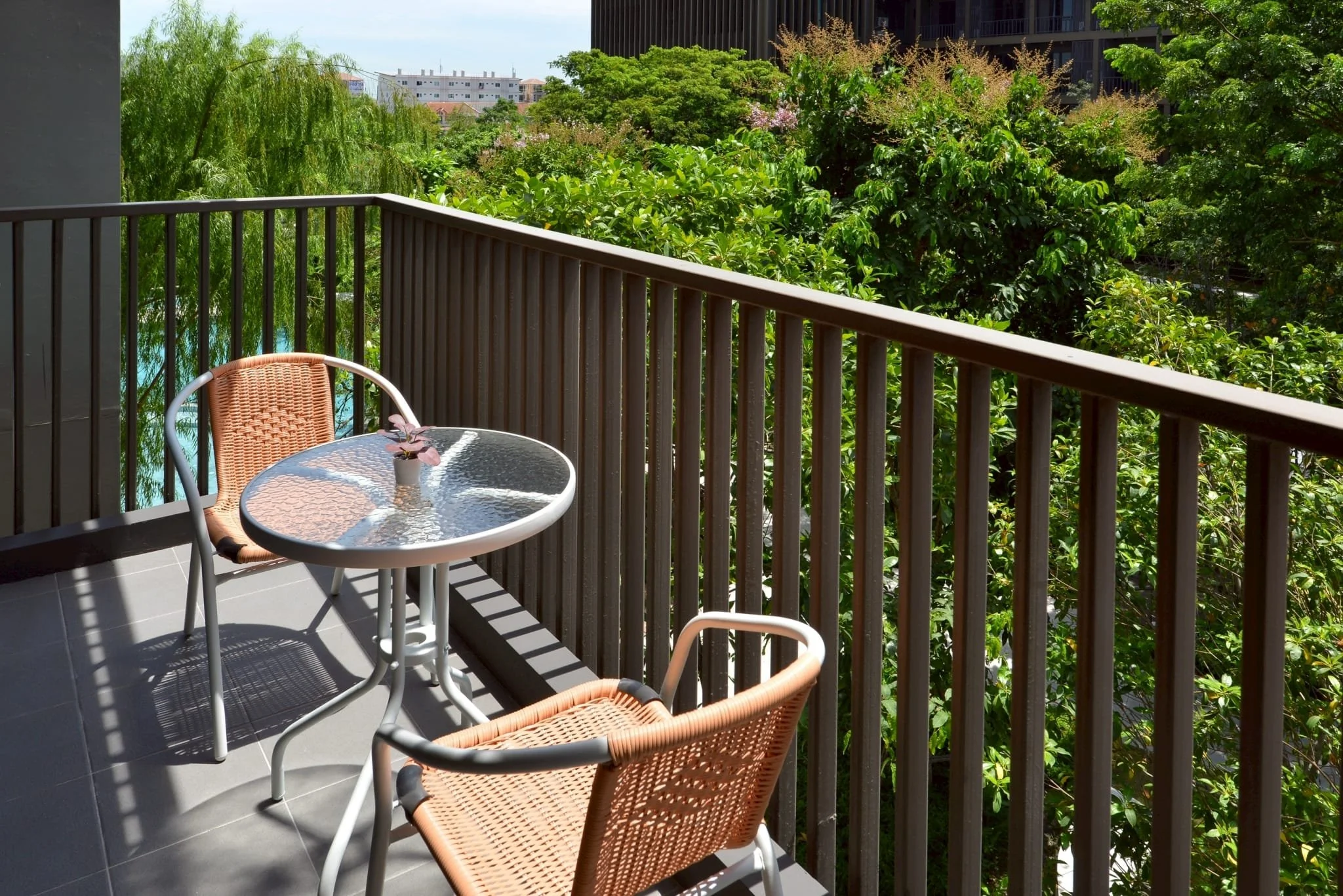 Balcony with a glass-top table, two woven chairs, greenery, and trees in an urban setting.