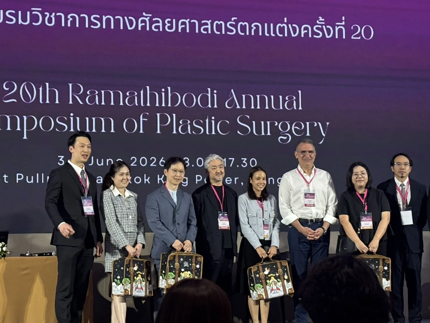 A group of eight people standing on a stage at the 20th Ramathibodi Annual Symposium of Plastic Surgery, with a large screen behind them displaying information about the event.