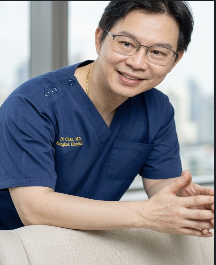 A man wearing glasses and a blue medical uniform with a name tag reading Dr.Dominic, Dr.Rushapol. He is smiling and sitting indoors near a window with a city view.