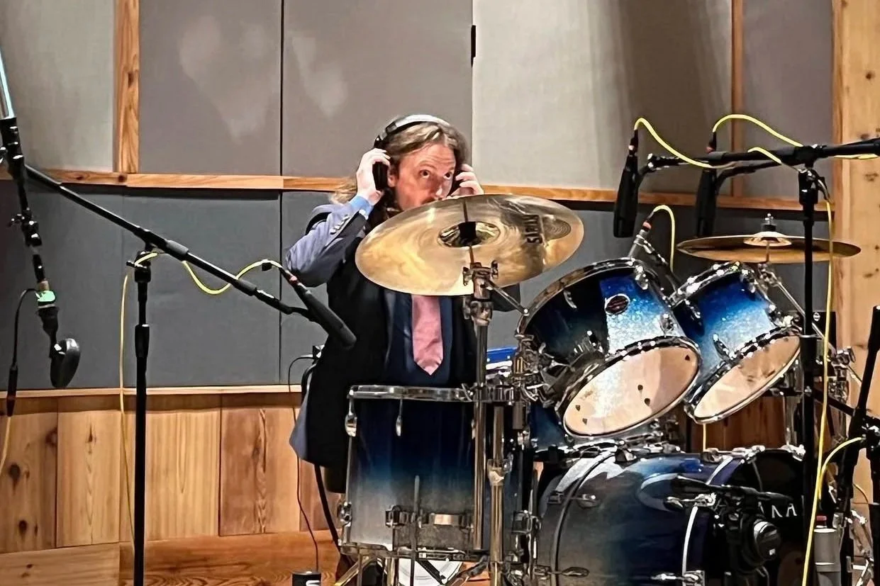 A woman in a recording studio, wearing headphones, sitting behind a drum set, appearing to be surprised or excited.