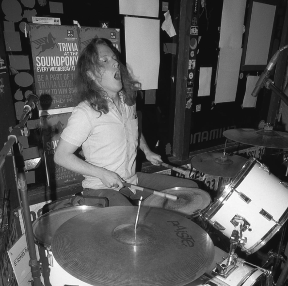 A woman passionately playing a drum set in a music venue with posters and flyers on the wall behind her.