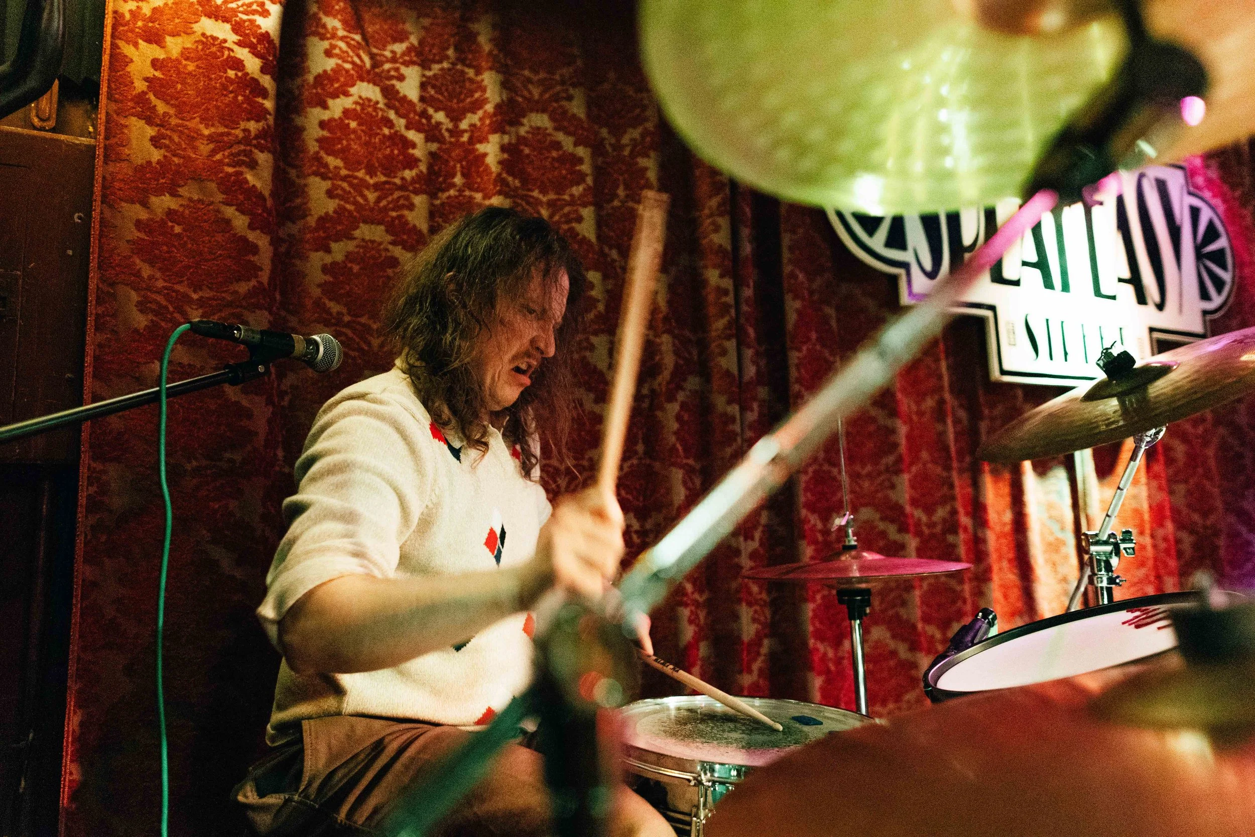 A musician with long hair playing drums on a stage with red and gold patterned curtains in the background.