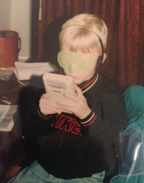 A young boy sitting and looking at a handheld gaming device, with green headphones and a face mask pulled up over his eyes, in a room with red curtains.
