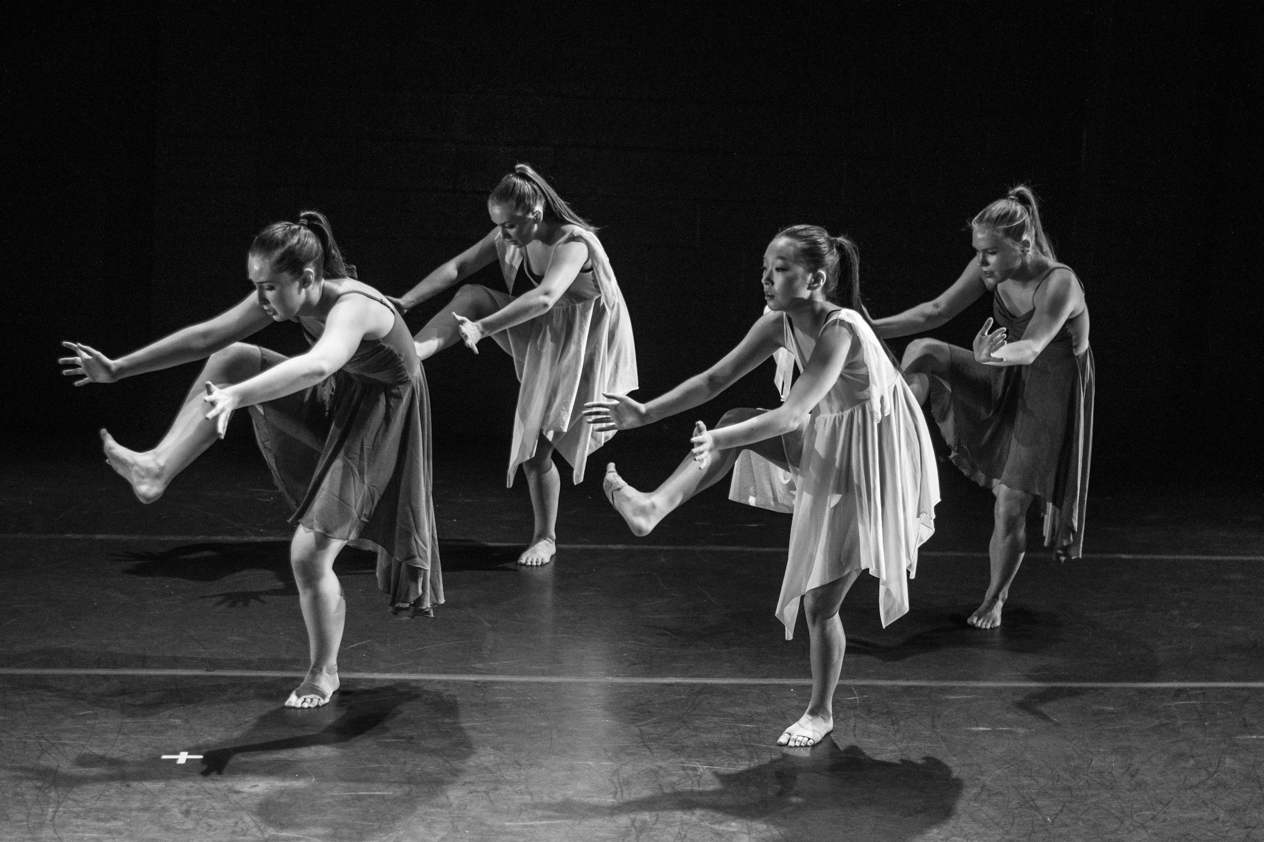 Black and white photo of four female dancers in a line on stage, performing a dance routine with arms extended forward and legs raised. They are dressed in sleeveless tops and flowing skirts, with their hair tied back, and are barefoot. The background is dark, focusing on the dancers.