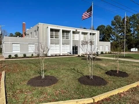 A white two-story building with a flagpole flying the American flag in front, surrounded by a grassy area and three young trees with mulch around their bases.