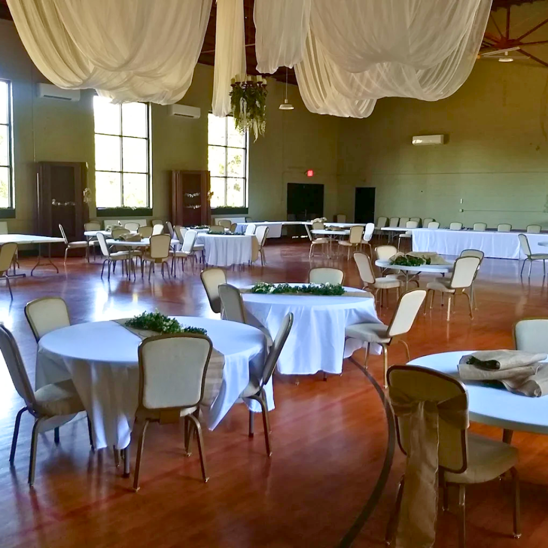 An event hall decorated with round tables covered in white tablecloths and surrounded by beige chairs, with large windows allowing natural light, and white drapery hanging from the ceiling.