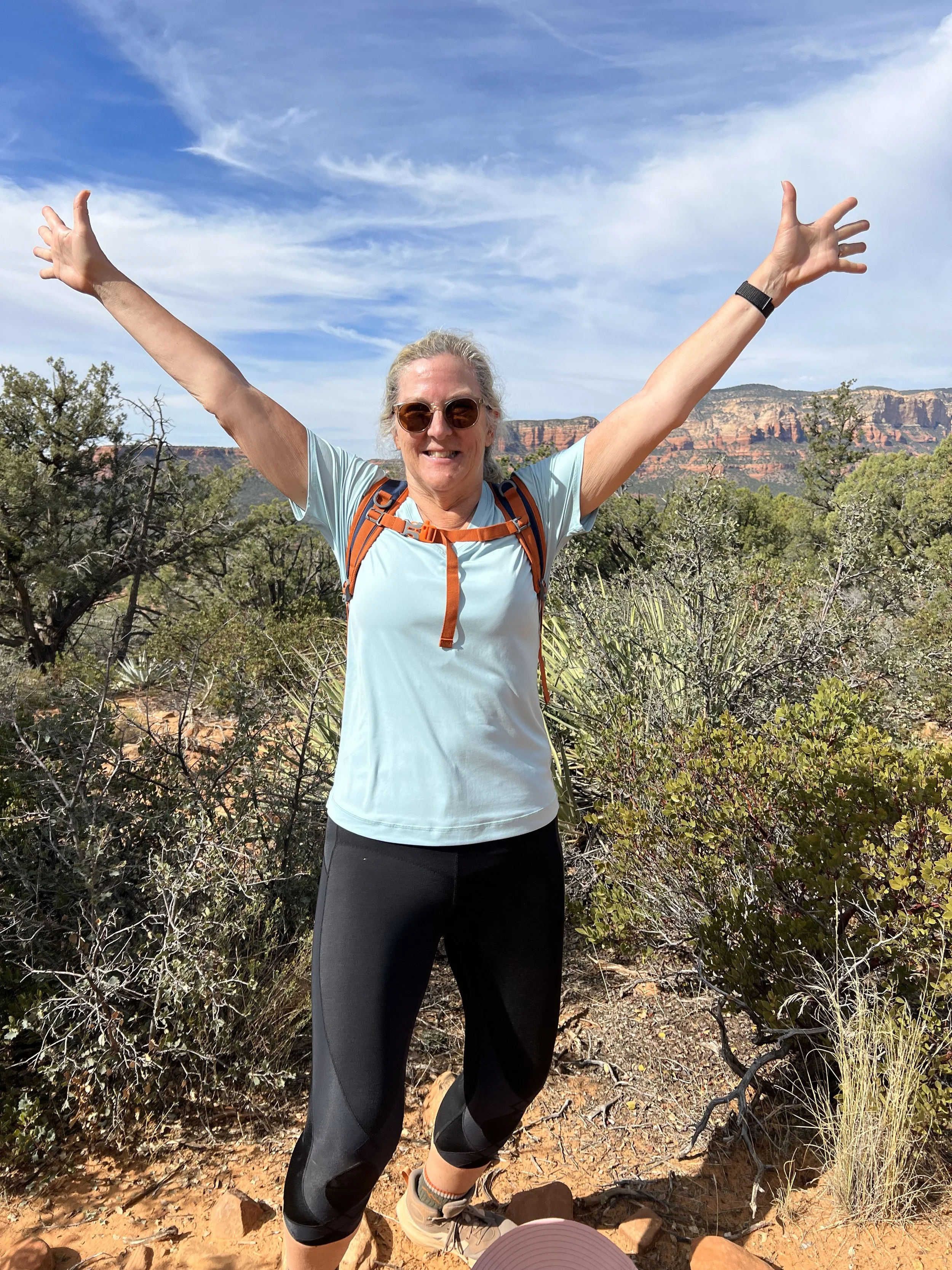 A woman with sunglasses, a light blue athletic shirt, and black leggings smiling with arms raised in a desert landscape with red cliffs and a blue sky.