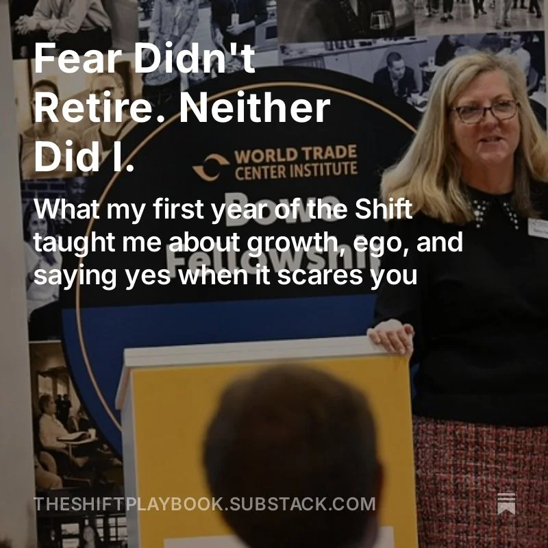 A woman with blonde hair and glasses standing in front of a display at the World Trade Center Institute. There are photographs of people in the background. The woman is smiling and wearing a black top with glasses.