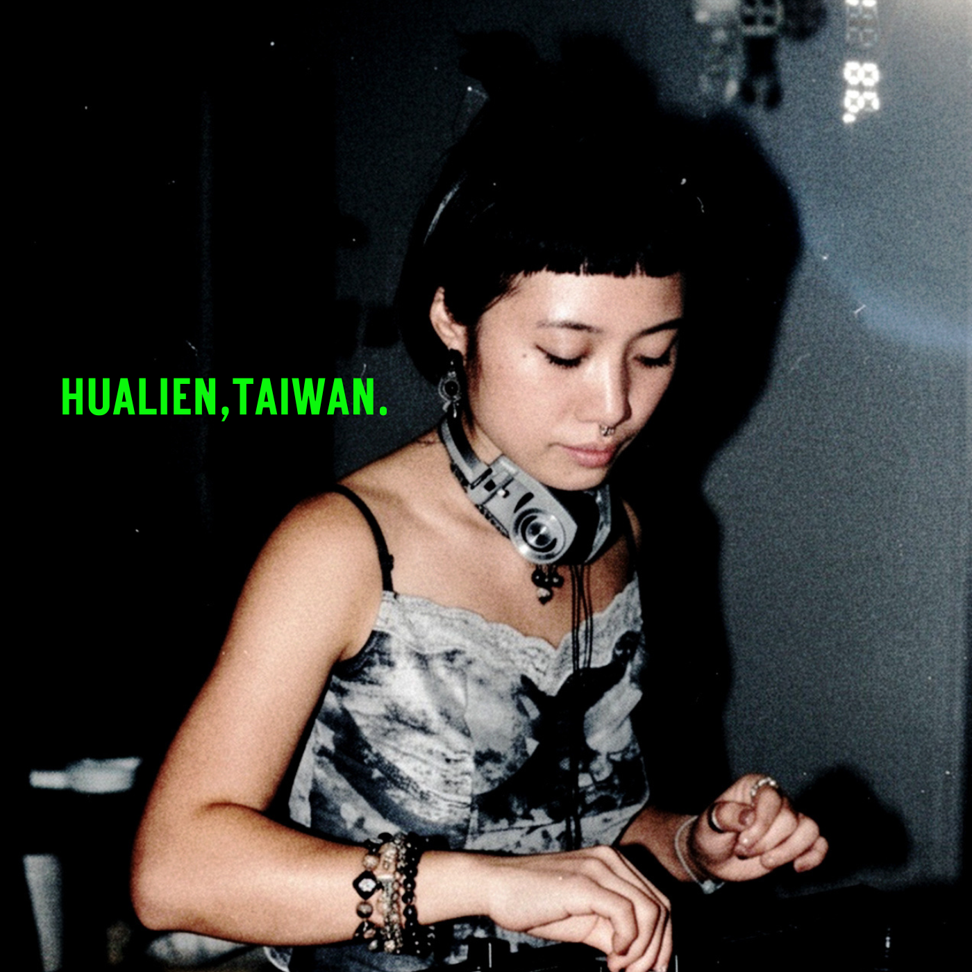 A young woman with short black hair and side-swept bangs, wearing headphones around her neck, a patterned sleeveless top, and layered bracelets, with her eyes closed, in a dimly lit room, with green text on the image reading 'HUALIEN, TAIWAN.'