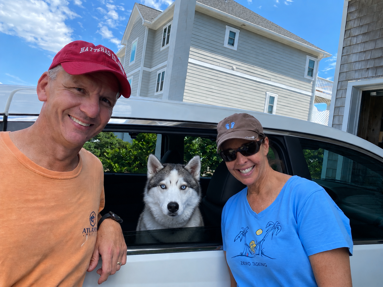 Two smiling people, a man and a woman, with a husky dog in a car's open window outdoors on a sunny day, with a residential house in the background.