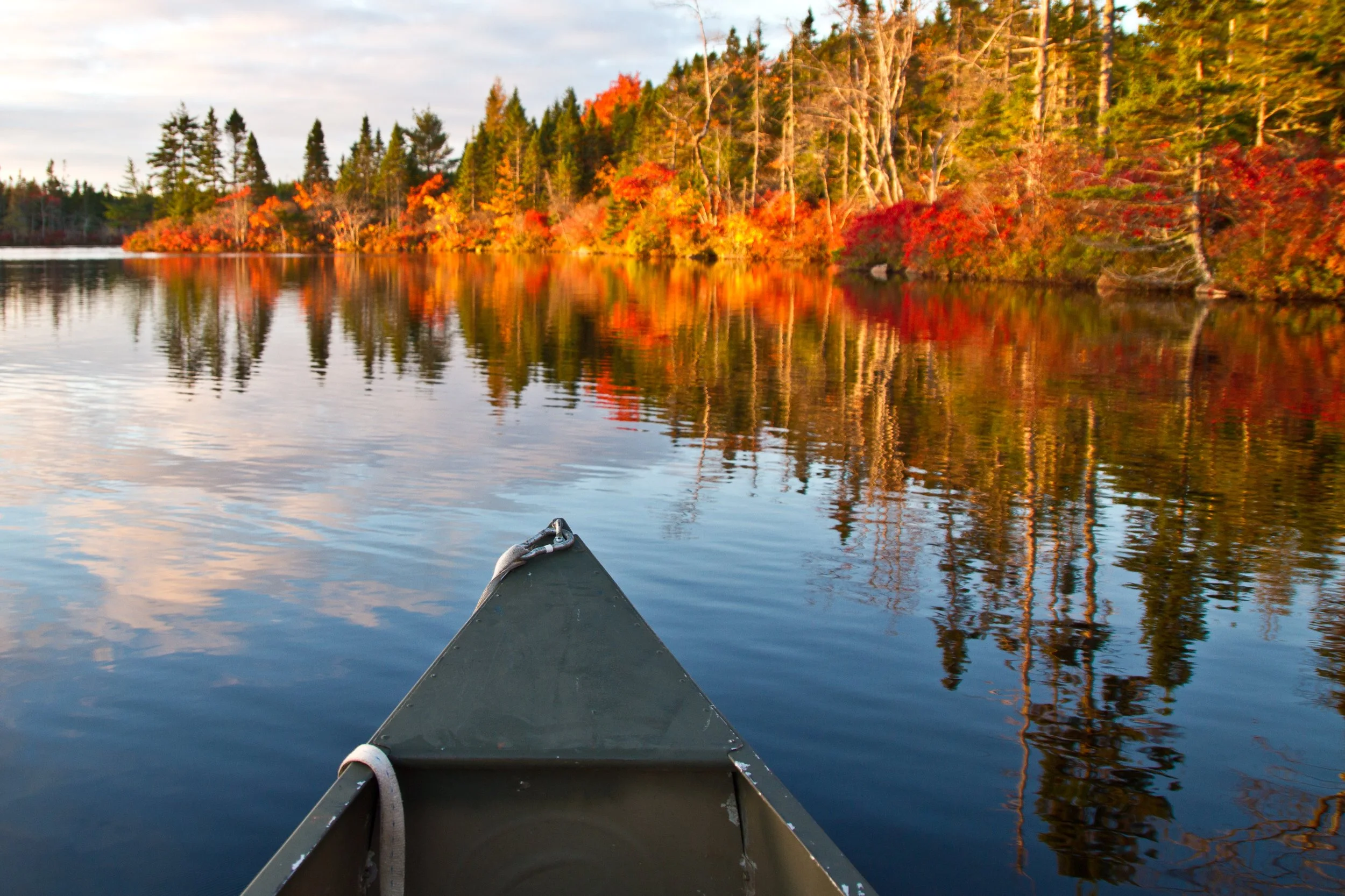 A canoe on a calm lake with vibrant autumn trees reflecting on the water, and a cloudy sky in the background.