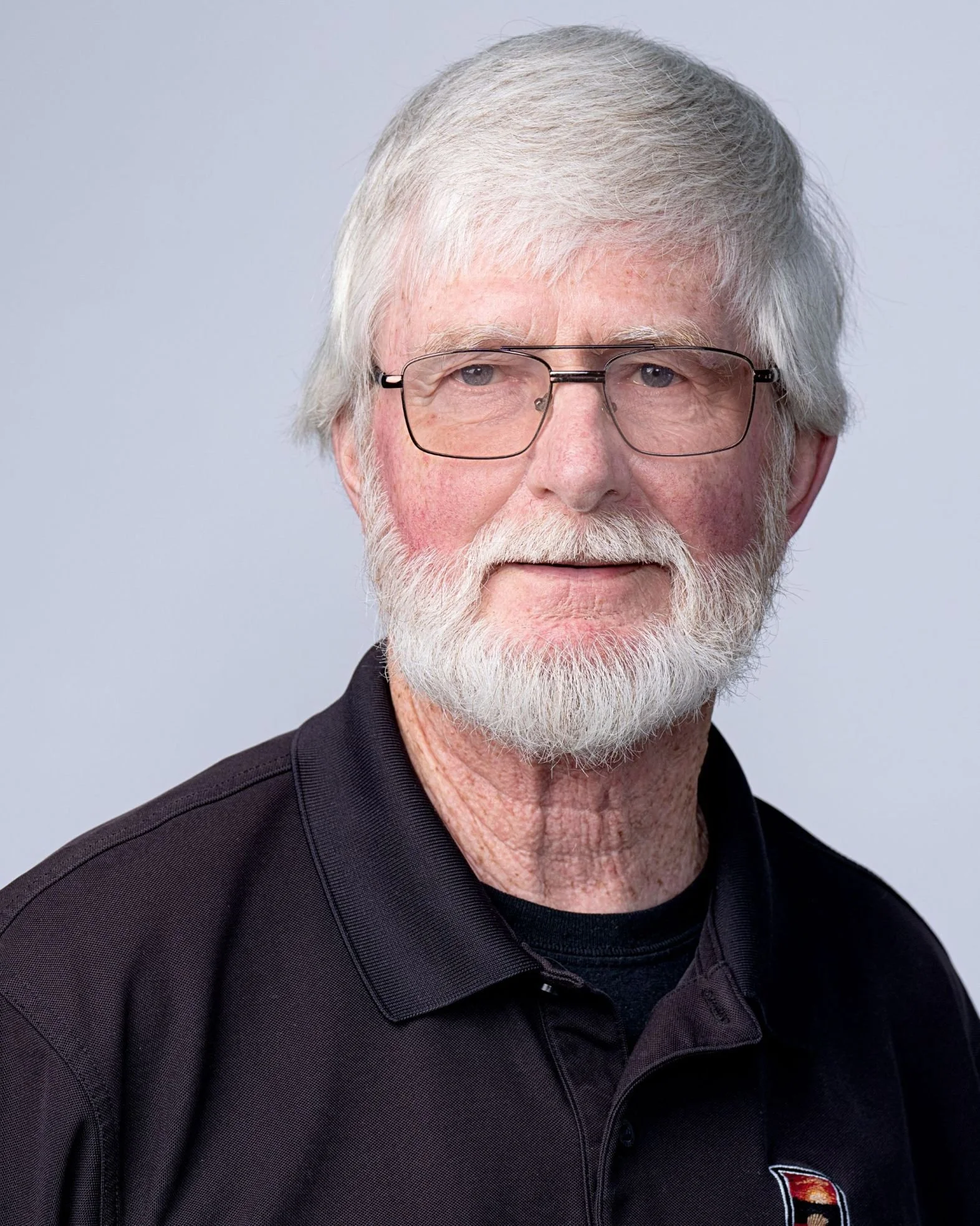 Close-up of an older man with white hair, glasses, and a beard, wearing a black collared shirt with a small logo on the chest, against a light gray background.