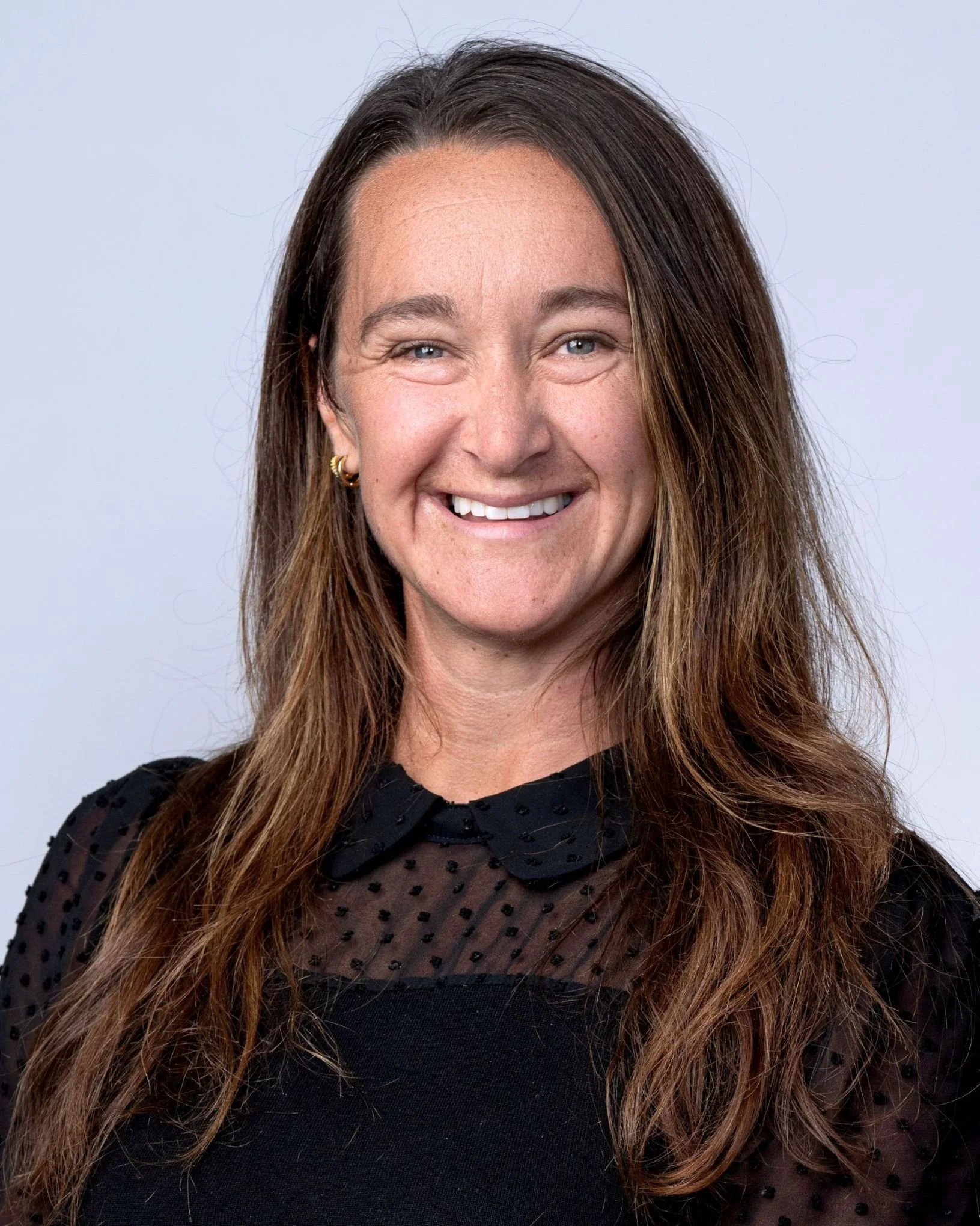 Close-up image of a woman smiling, with long wavy brown hair, wearing a black top with tulle sleeves, and small gold hoop earrings, against a plain light background.