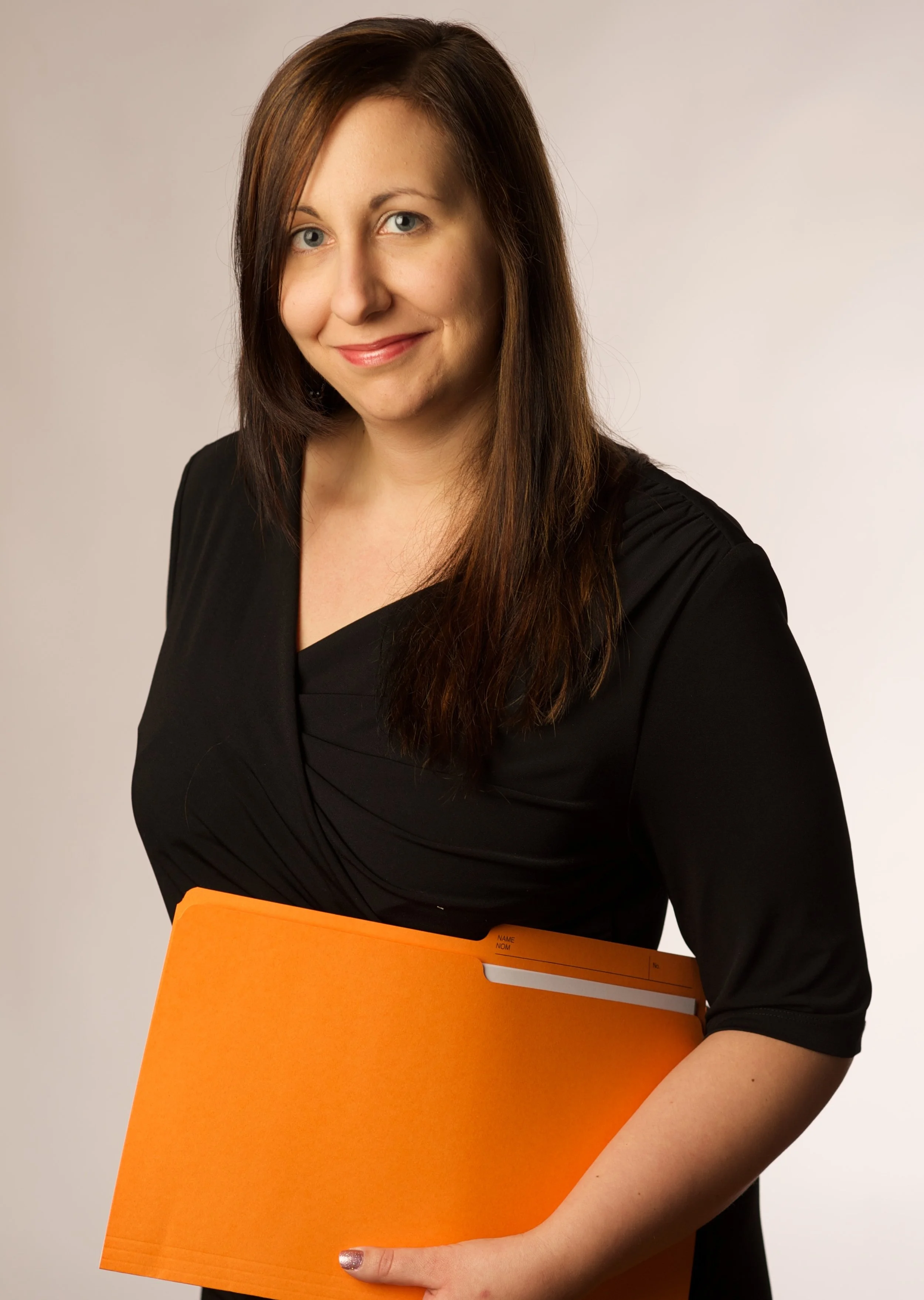 A woman with shoulder-length brown hair and blue eyes smiling, wearing a black top, holding an orange folder. The background is light gray.