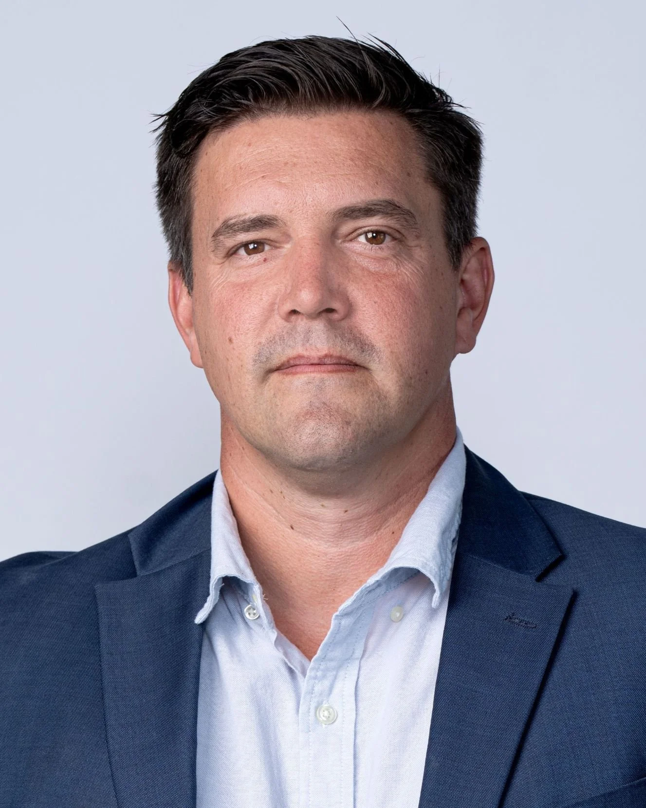 A professional man with dark hair, wearing a dark blue suit and a light blue collared shirt, standing against a plain light background.