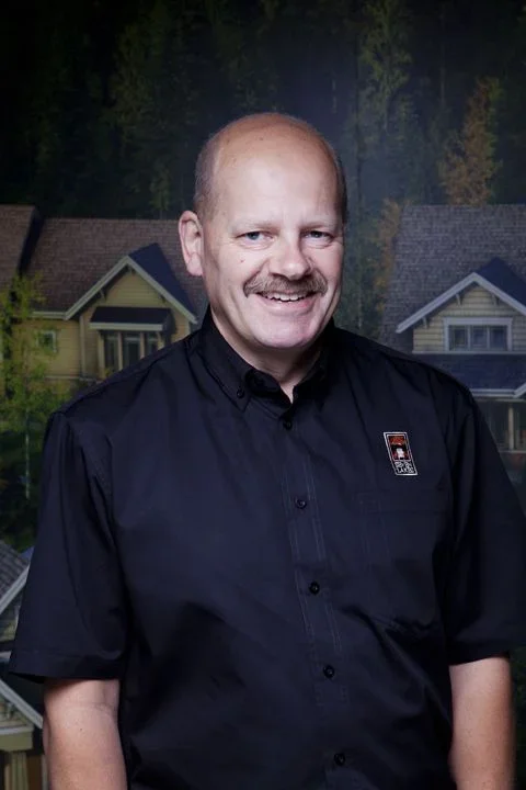 A man with a bald head, mustache, and light skin, wearing a black shirt with a logo, smiling in front of a backdrop of houses and trees.