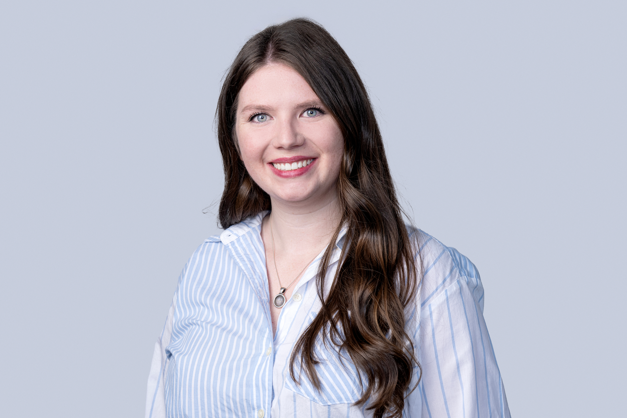 A young woman with long, wavy brown hair and blue eyes, smiling, wearing a light blue and white striped shirt, against a plain light gray background.