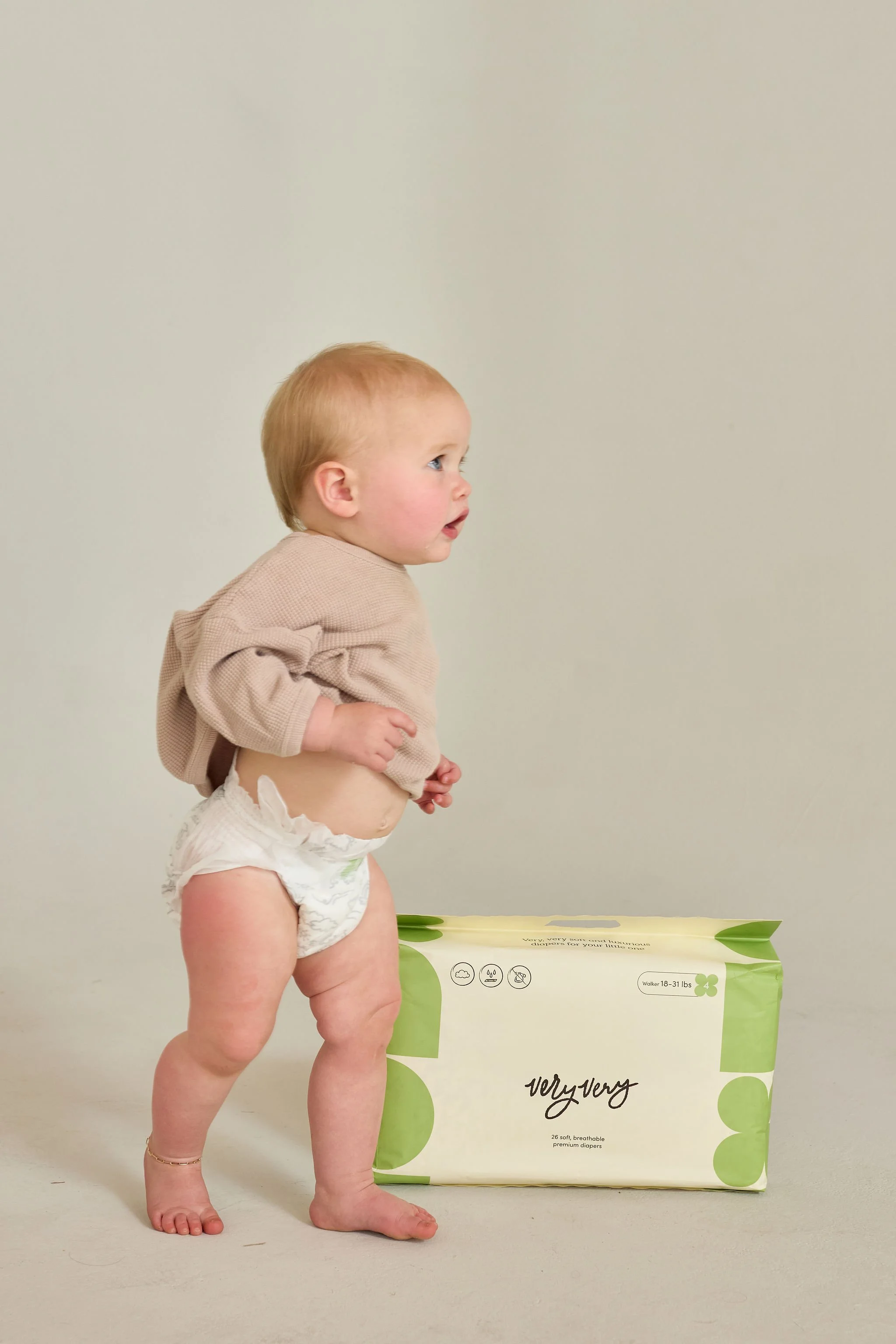 A baby standing next to a pack of diapers on a plain background.
