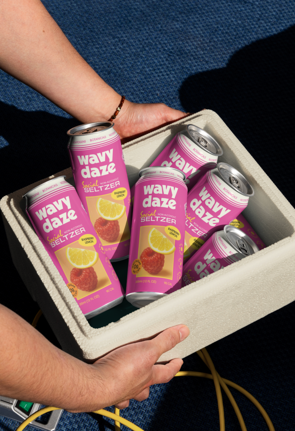 A person's hand holding a white insulated cooler filled with pink cans of Wavy Daze Raspberry Lemon flavored non-alcoholic social seltzer.