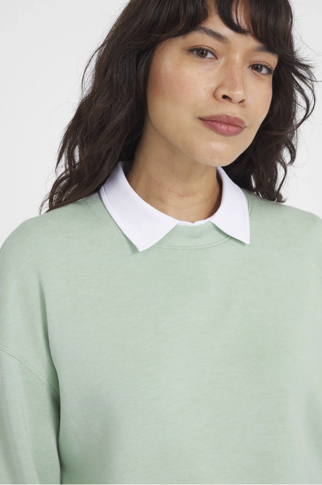 A woman with wavy brown hair wearing a light green sweatshirt with a white collared shirt underneath.