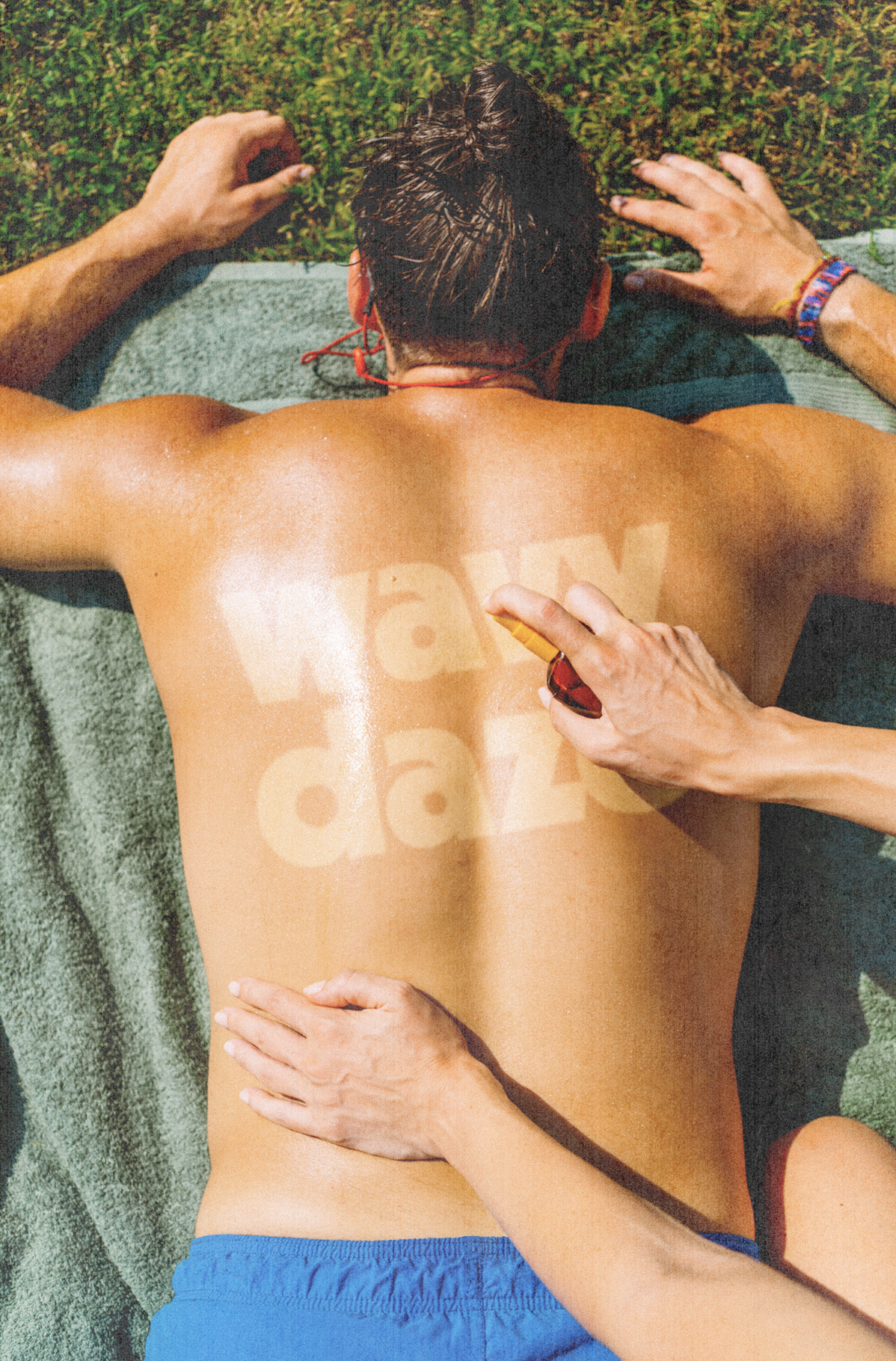 A person lying face down on a towel on the grass, getting a tanning session while someone applies sunscreen on their back.