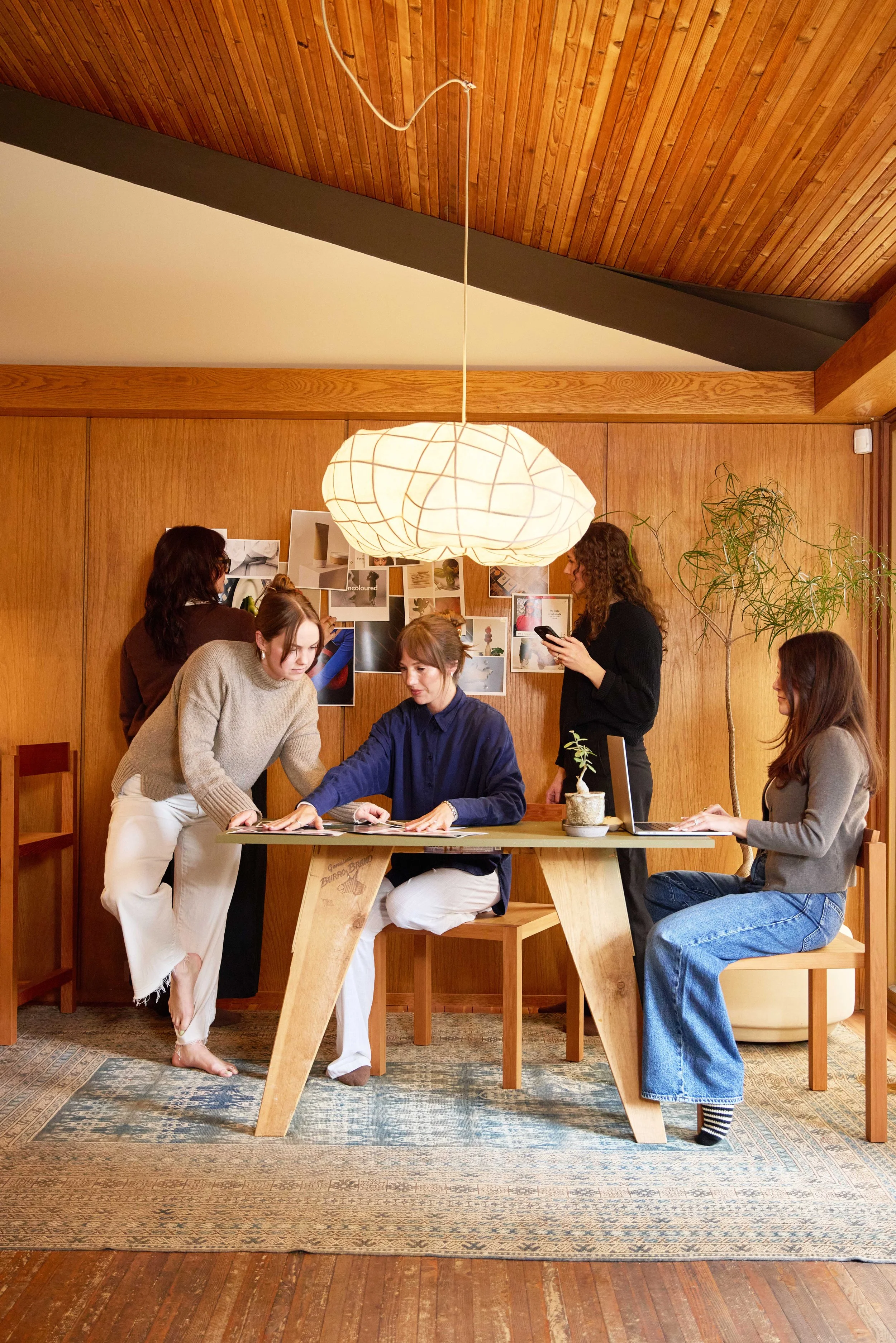 Five women working together in a cozy wooden room, with one seated at a table with a laptop, two standing, one looking at a phone, and one sitting at the table, in front of a wall with photos and mood boards, and a large hanging light fixture above.