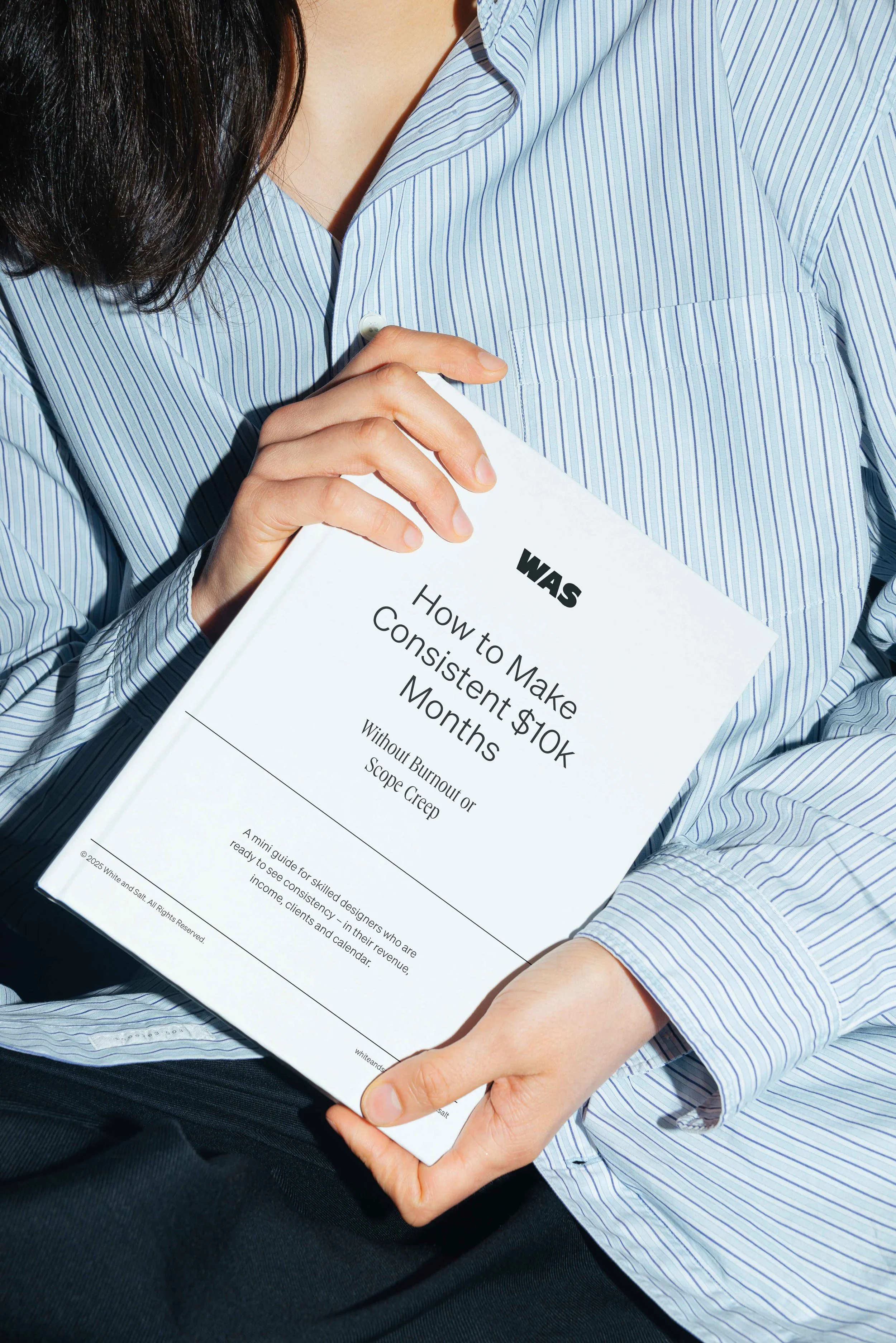 Person holding a white book titled 'How to Make Consistent $10K Months Without Burnout or Scope Creep'