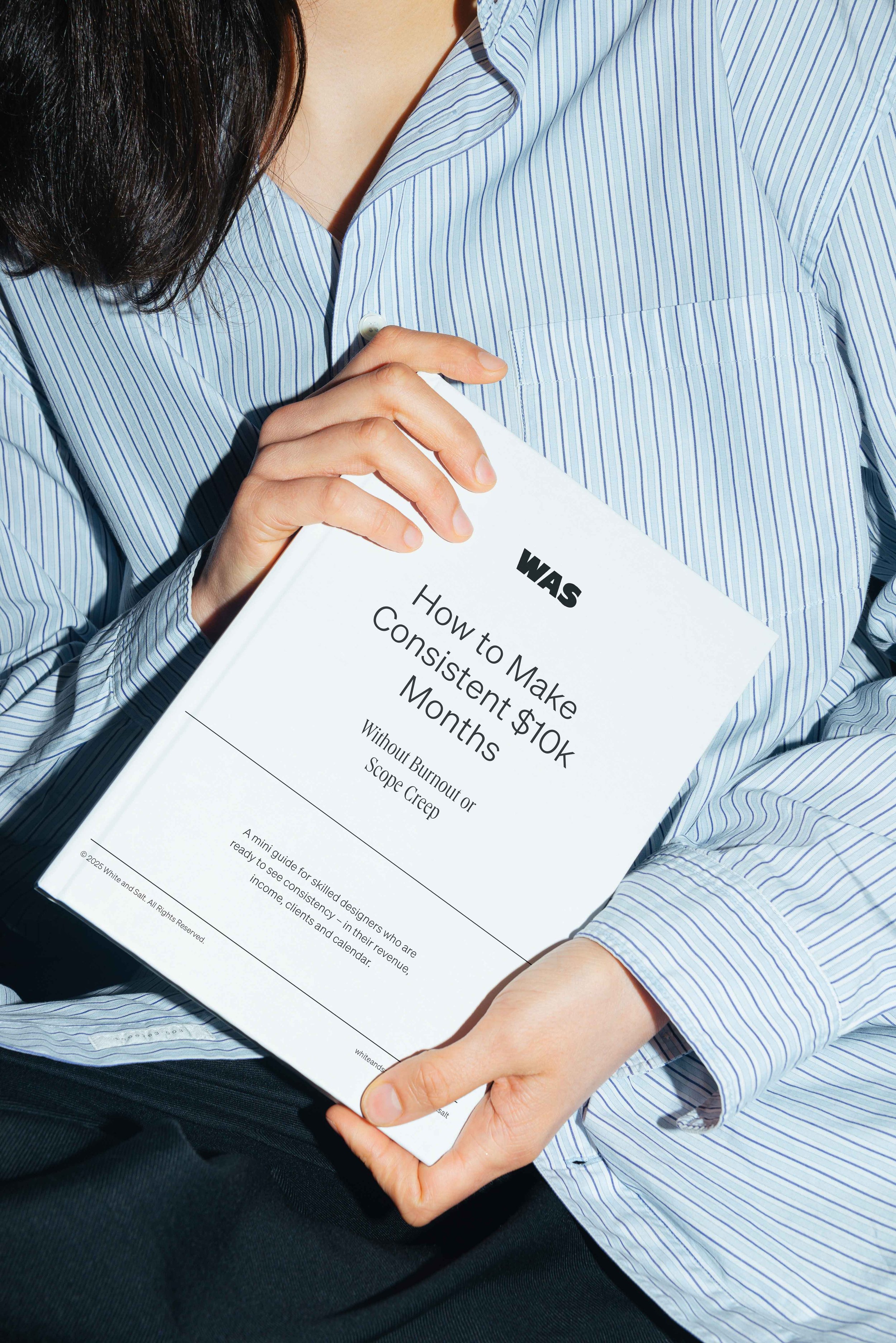 Person holding a book titled 'WAS How to Make Consistent $10K Months Without Burnout or Scope Creep'