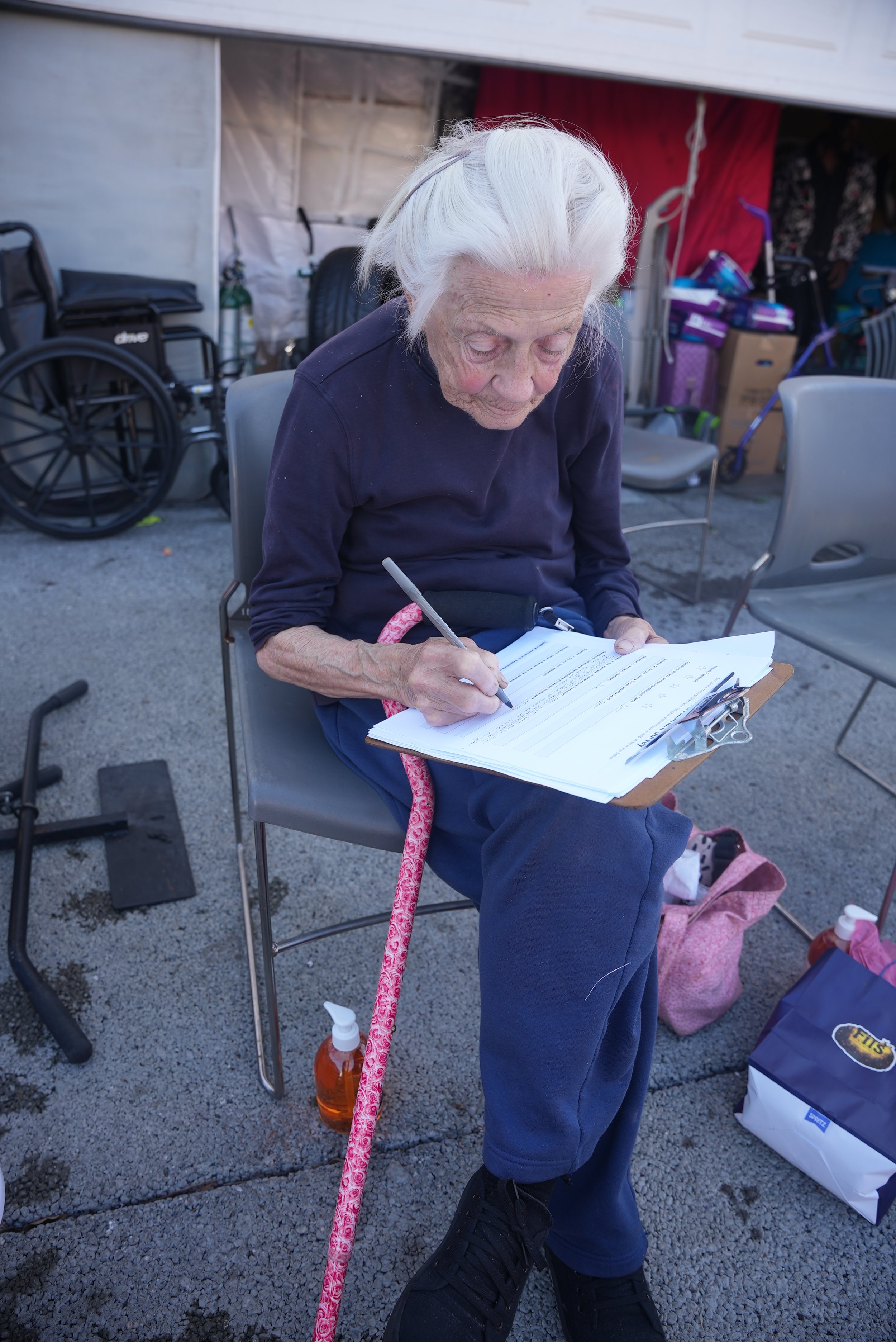An elderly woman with white hair, wearing a dark long-sleeved shirt and pants, sits outdoors on a folding chair, writing on a clipboard with papers. She has a pink walking cane with floral design leaning beside her. A bottle of hand sanitizer is nearby. In the background, there are chairs, a wheelchair, and various supplies against a structure with fabric and boxes.