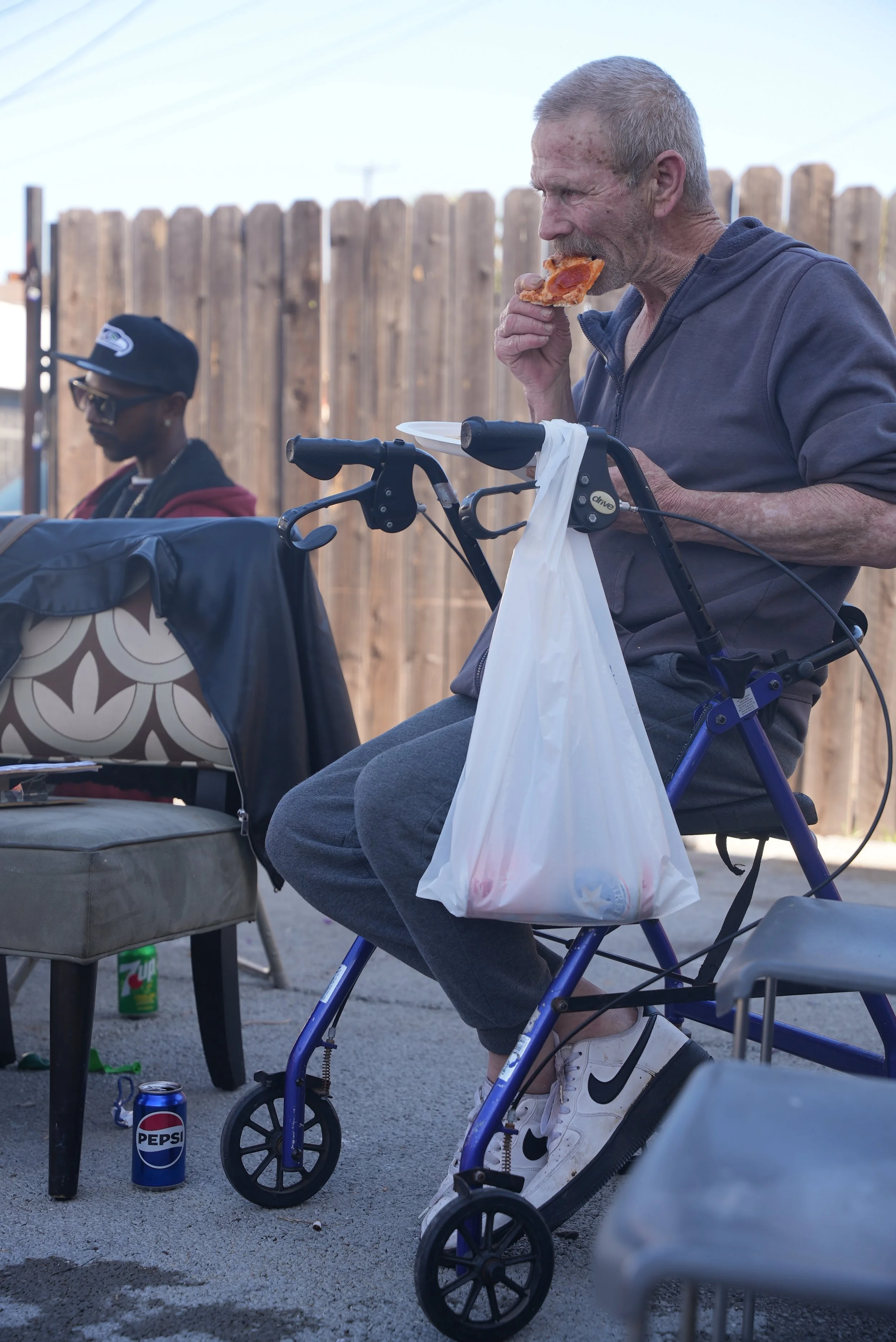 An elderly man in a wheelchair eating a slice of pizza, with a plastic bag hanging on the wheelchair handle, and a young person in the background wearing sunglasses and a cap. There is a Pepsi can, a soda can, and a chair nearby in an outdoor setting with a wooden fence.