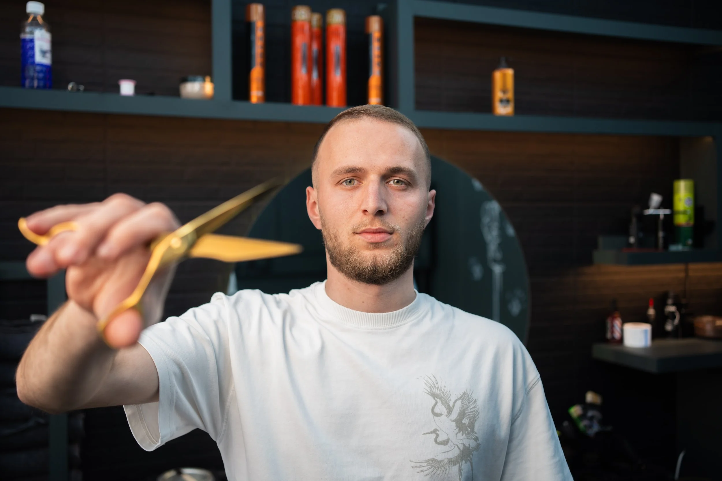 A young man with short hair and a beard is holding scissors, preparing to cut hair or beard. He is standing in a barbershop or salon with shelves of hair products behind him.