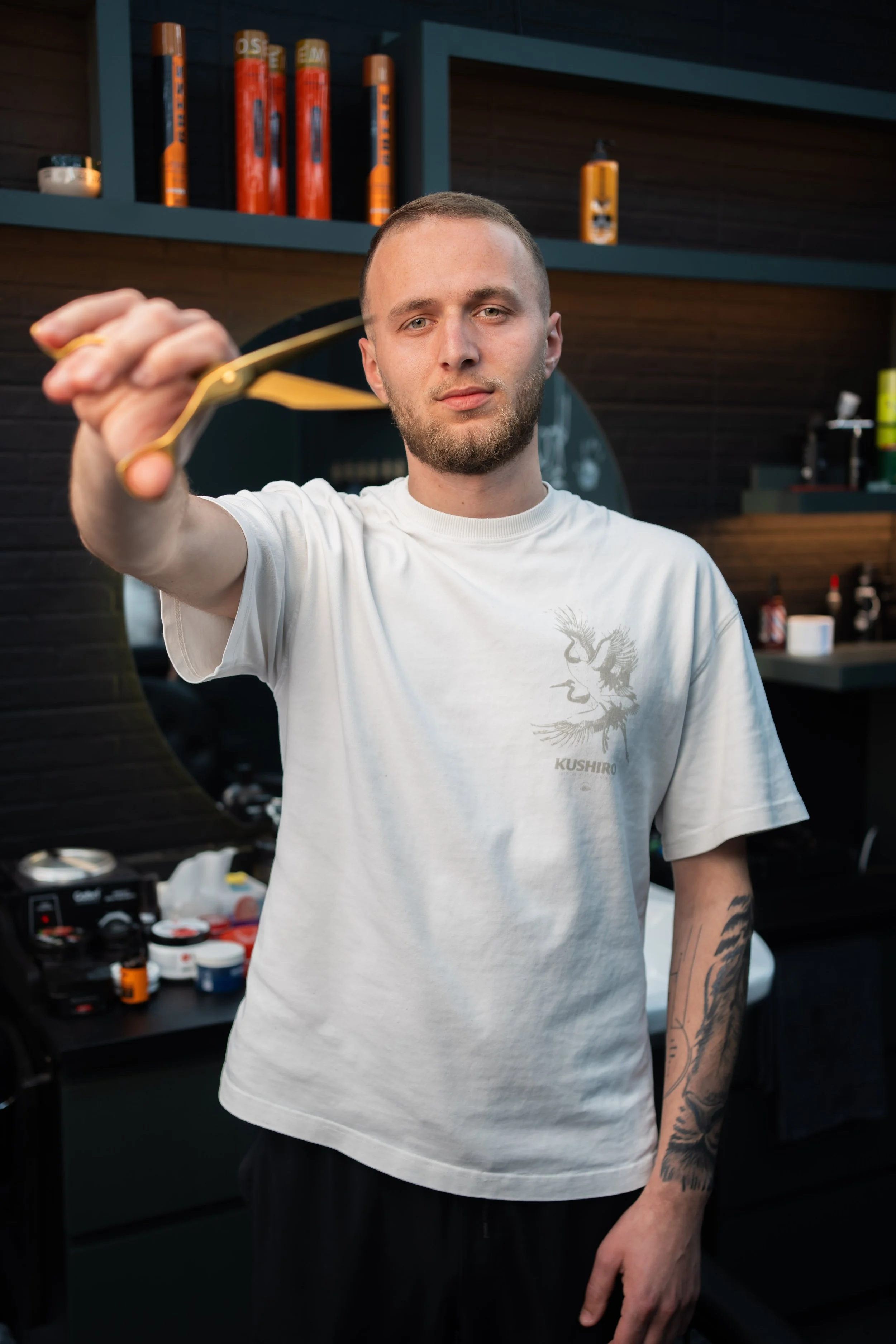 A man with short hair and tattoos on his arm is holding a gold-colored object towards the camera. He is wearing a white T-shirt with a dragon design and the word 'KUSHIRO' on it. The background features a dark kitchen with shelves holding various bottles and jars.