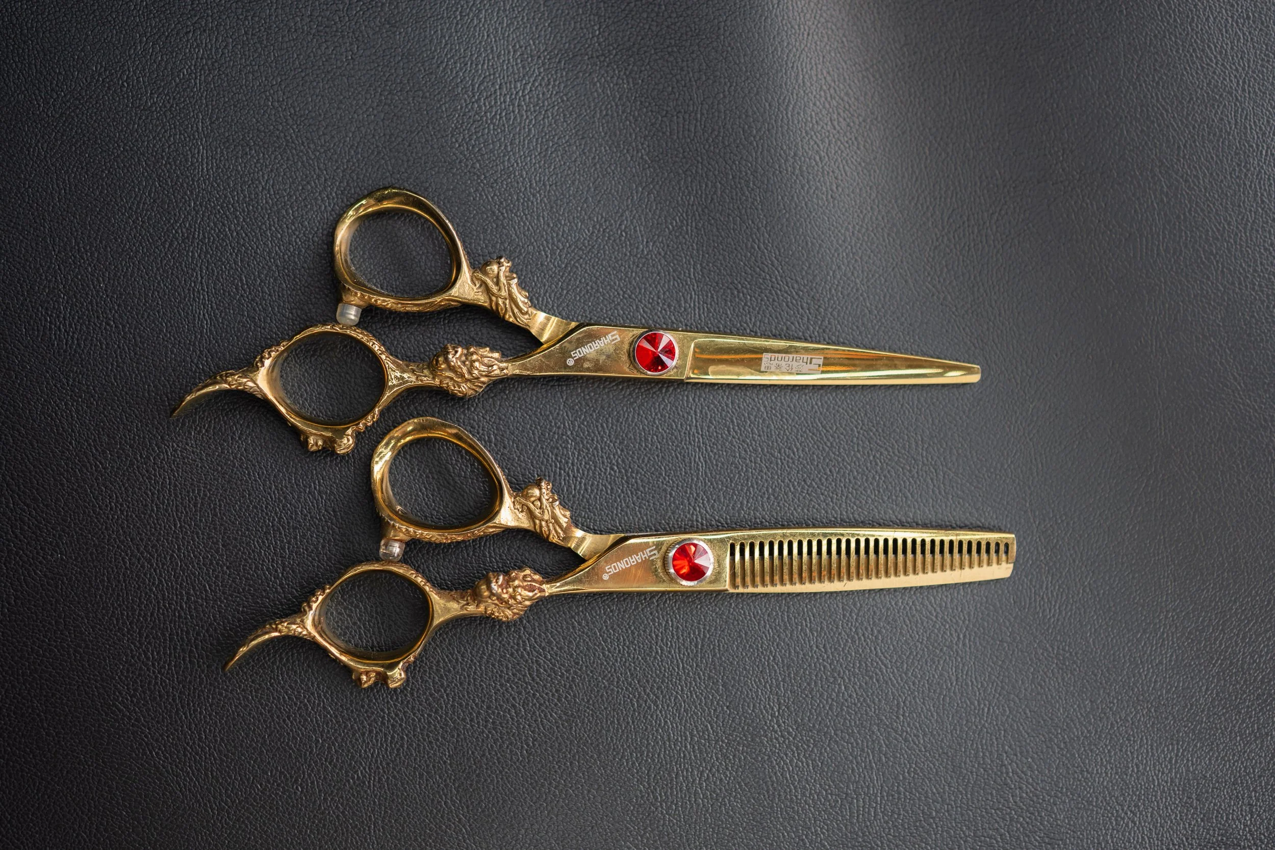 A set of three gold-colored hair cutting scissors with decorative handles and a red gem accent, placed on a black textured surface.