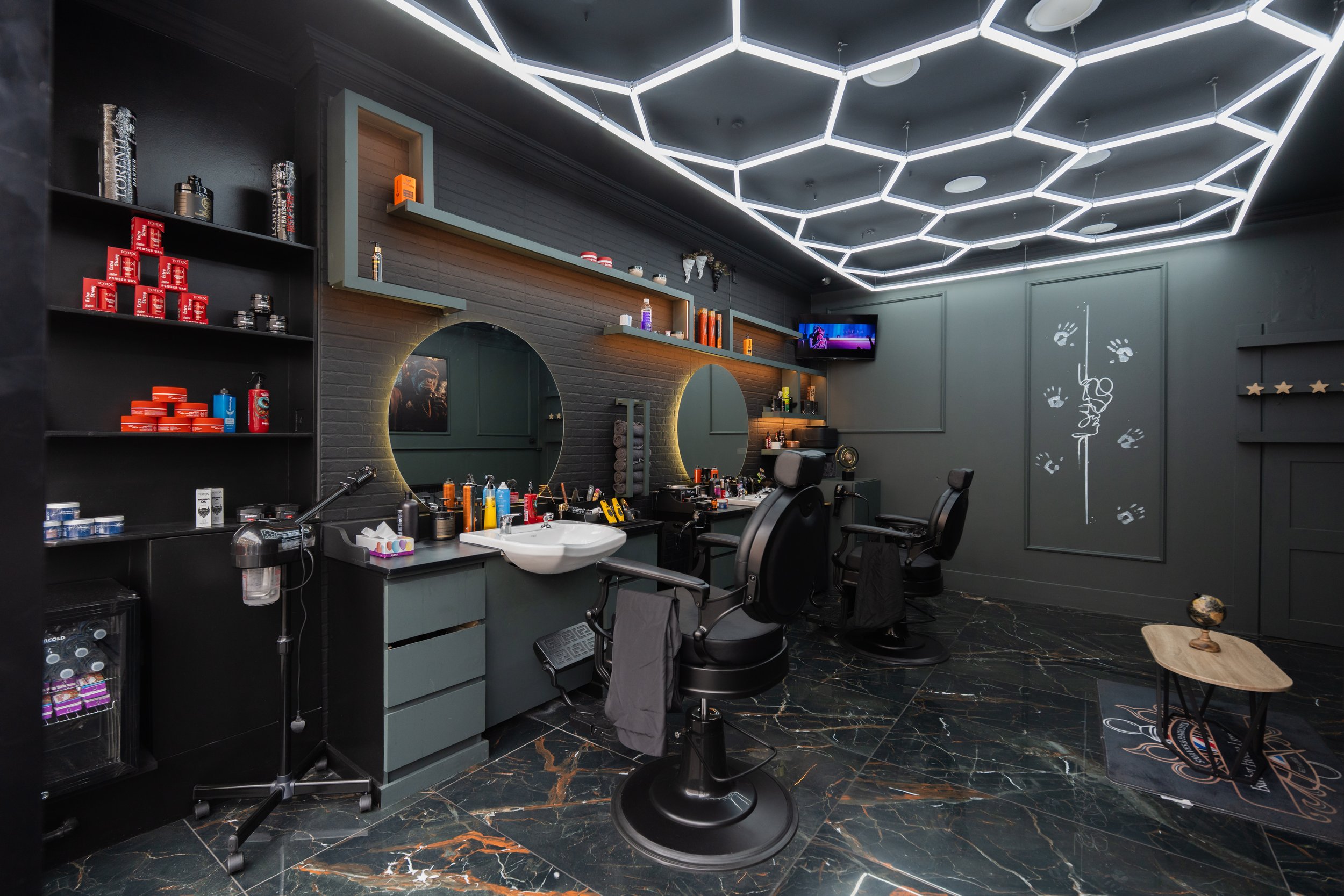 Modern hair salon with black shelves holding haircare products, two round mirrors on a gray wall, and three barber chairs in front of stations with styling tools. Unique hexagonal LED ceiling lights, a black marble floor with orange veins, and wall art including a handprint with hanging scissors and a TV screen in the corner.