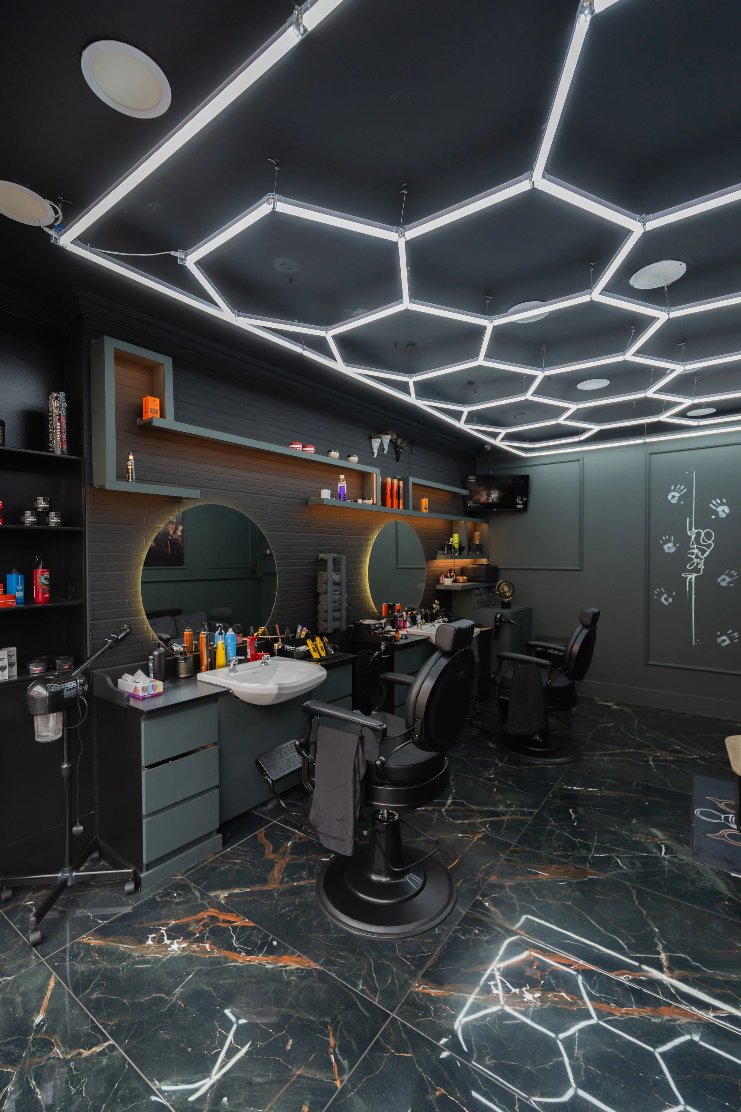 Interior of a modern barbershop or hair salon with black and gray decor, illuminated hexagonal ceiling lights, black chairs, and marble flooring with orange and white veining.